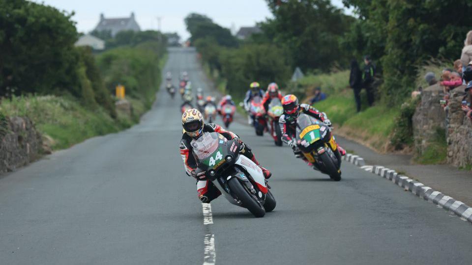 Lightweight machines on the Billown Course, which is flanked by hedgerows with some spectators seen to one side, with the leaders leaning into a right-hand bend.