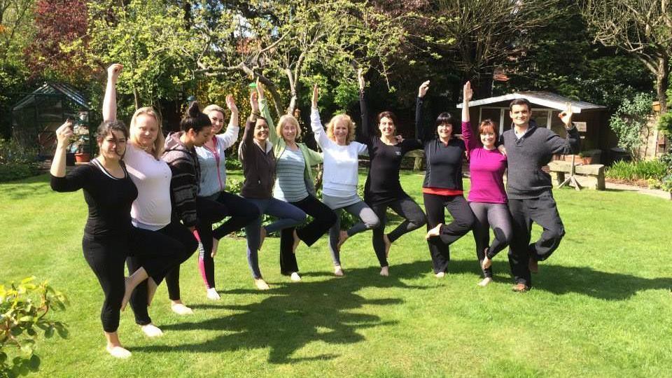 Eleven people stand in a garden doing yoga poses.