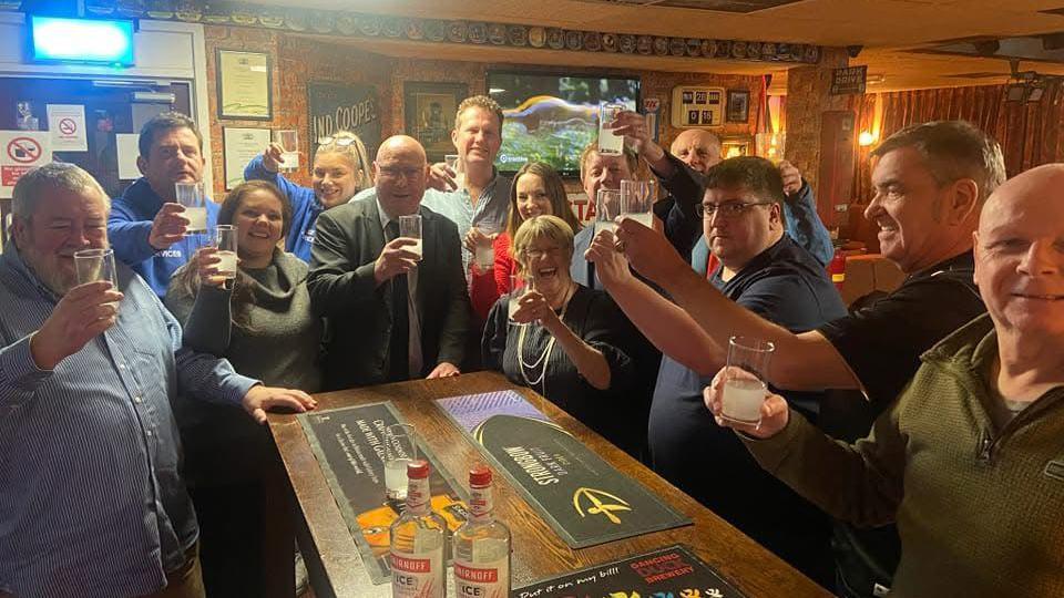 A group of people stand around a pub bar all raising their glasses as they make a toast.