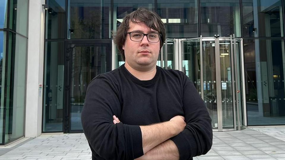 Councillor Paul Edgeworth standing in front of Sunderland's City Hall. He has brown hair and is wearing glasses and a black long-sleeve jumper. He is standing with his arms crossed.