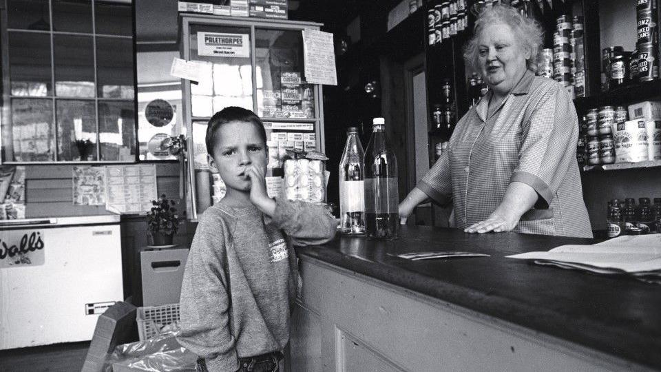 Llun du a gwyn o siop ym Mhontrhydygroes. Mae bachgen yn edrych ar y camera tra'n sefyll wrth gownter y siop gyda'i law dde yn ei boced. Mae menyw tu ol y cownter yn edrych lawr ar y bachgen. Mae dwy botel wydr ar y cownter a nwyddau'r siop ar silffoedd yn y cefndir ac ochr y llun.   