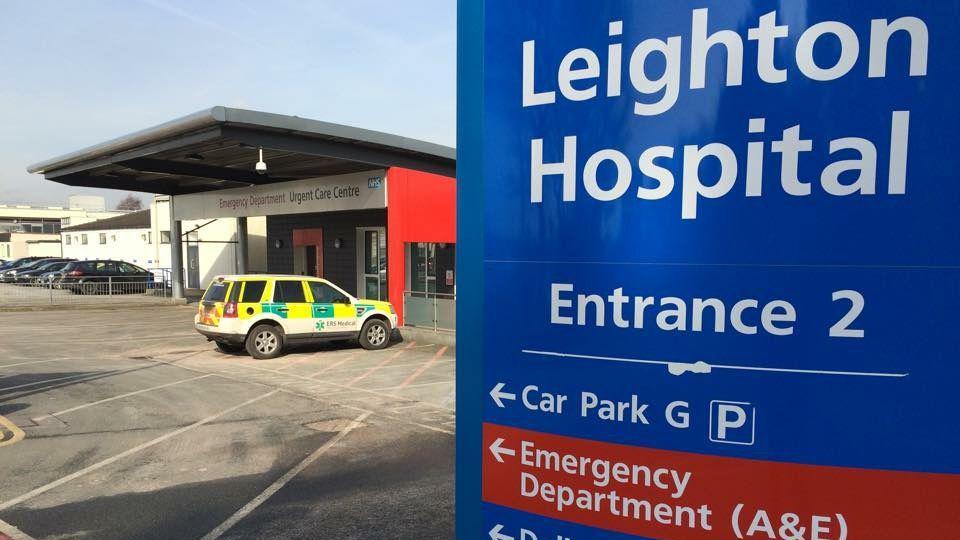 The Emergency Urgent Care Centre at Leighton Hospital with a blue sign in the front of the frame outside that reads 'Entrance 2'. It has arrows pointing towards the car park and A&E. A medical vehicle is parked outside.