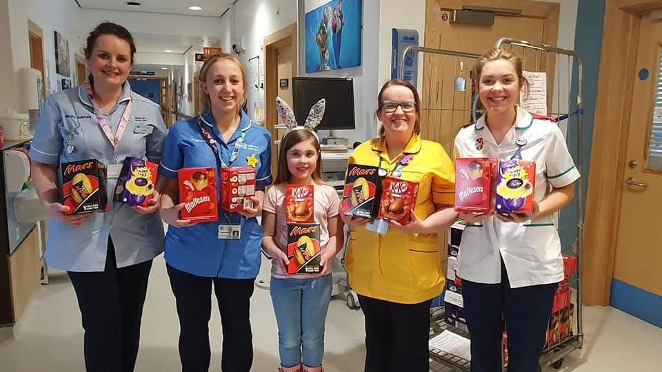 A little girl is standing beside four hospital staff members. They are all holding Easter eggs and smiling at the camera. The background is a hospital corridor with white walls and wooden doors. 