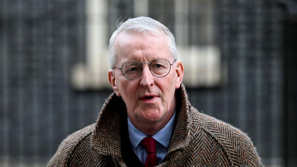 Hilary Benn with short grey hair and black glasses wears a tweed jacket with a blue shirt and red tie on beneath it.