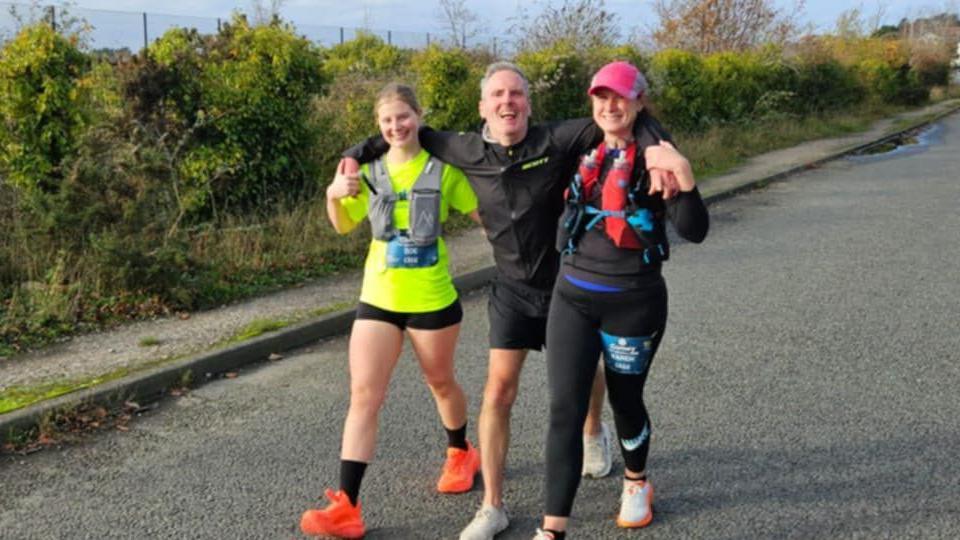 Mark running along a road towards the end of the half marathon - he's now being supported by two women with his arms around their shoulders, and the three of them are running together. All are wearing running kit.