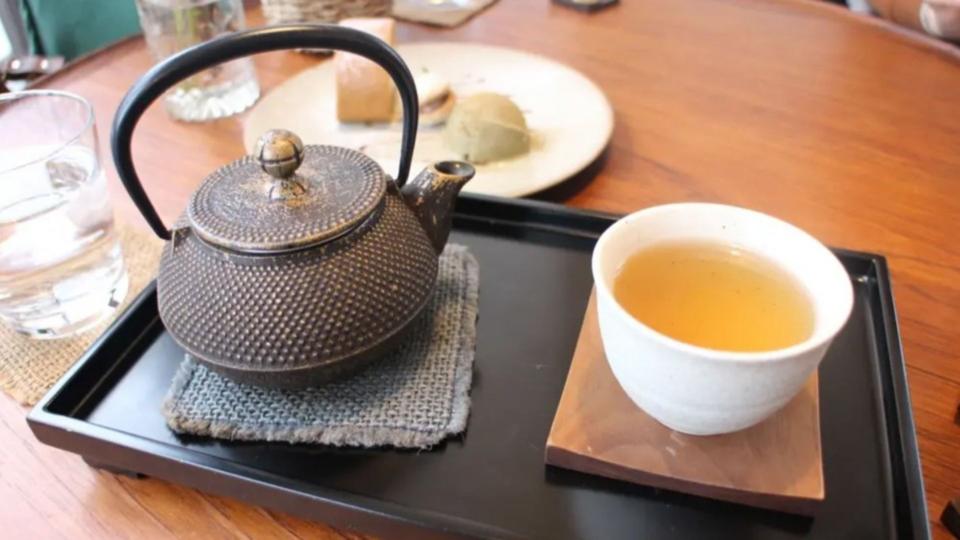 A brown teapot sits next to a white mug full of golden tea. Together they sit on a black tray on a brown wooden table