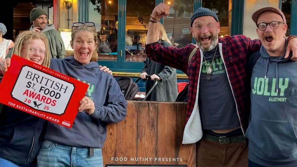 Four people, two men and two woman, clench their fists and cheer in celebration as they face the camera, They are standing either side of a wooden panel and the two women have a red and white placard with "British St Food Awards" on it.
