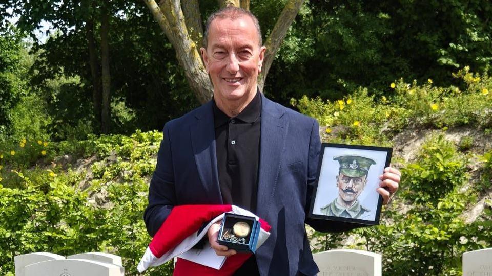 A smiling man in a suit holding a folded flag, pocket watch and drawing of a solider in a frame
