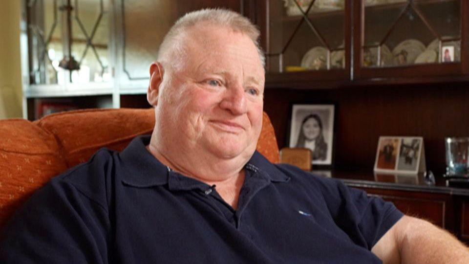 Ray, a 62-year-old man with closely cropped hair, smiling slightly as he looks towards the camera during an interview at his home.