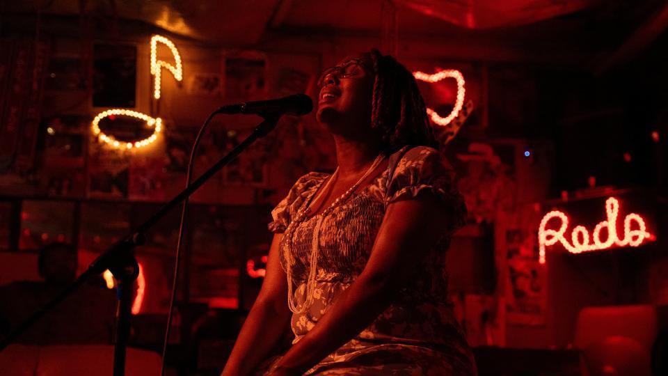 Bathed in red light, a woman sings into a microphone with her eyes closed