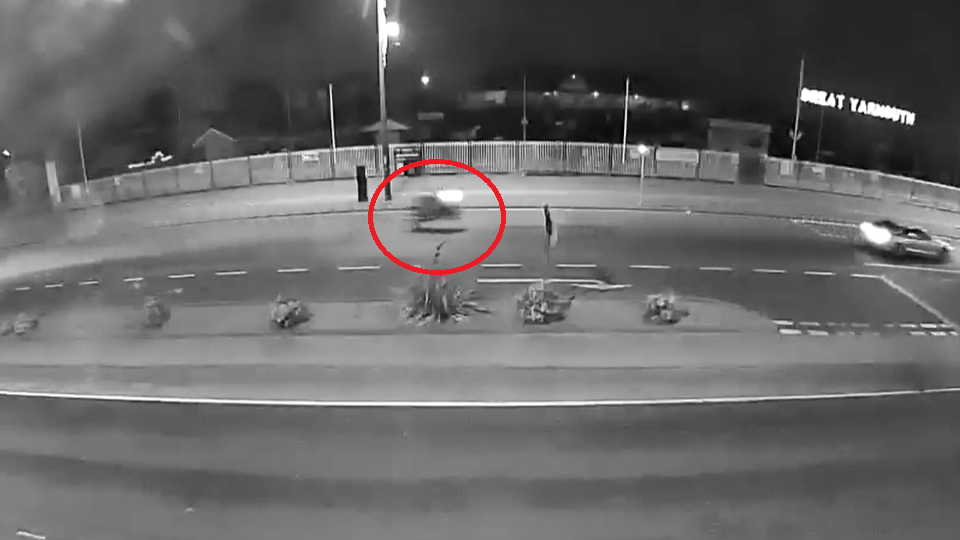 Black and white noise detection camera footage of Great Yarmouth seafront.  red circle highlights a motorbike with its lights on pulling a wheelie while travelling along the road 