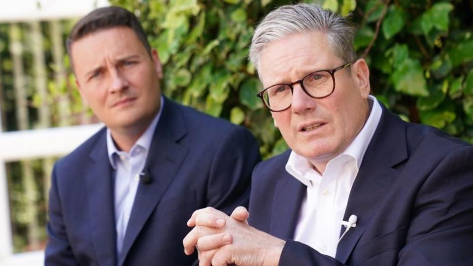 Wes Streeting on the left, Starmer on the right, with a green bush in the background