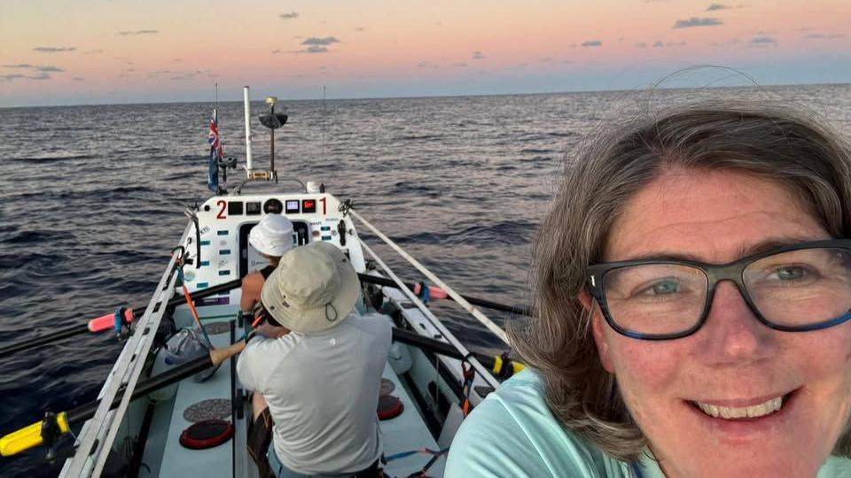 A woman with brown hair and glasses takes a selfie with two crew members in the background wearing hats while rowing.