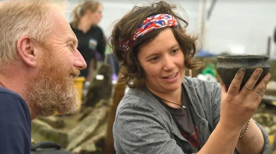 An archaeologist - a woman in a grey shirt and red and blue headband - holding an object that looks like a pot showing it to a man in a blue shirt and grey beard.