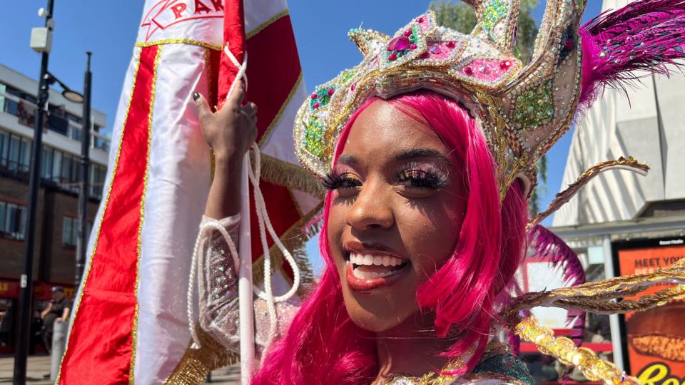 A woman wearing a carnival outfit complete with a pink wig and multi-coloured headpiece smiles at the camera. She holds a large flag in one of her hands.