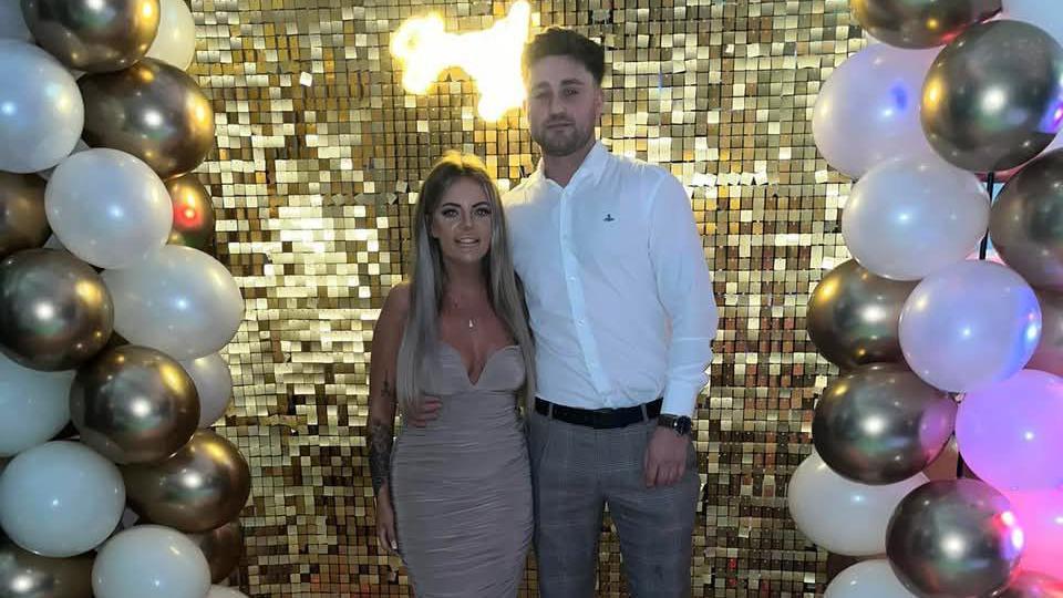 A young woman and man stand side by side, surrounded by a balloon arch, in front of a glittering gold background.