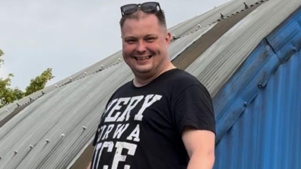 Tomasz Stankiewicz. He has short dark hair and no facial hair. He has black sunglasses on his head and is wearing a black t-shirt with white text on it. He is looking at the camera and smiling 