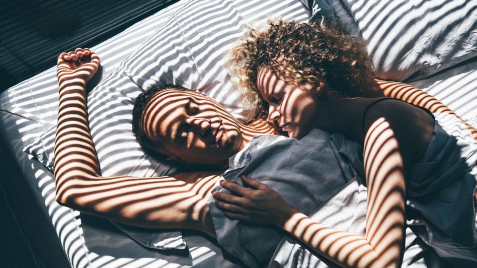 A man and a woman lay in bed cuddling. There's light shining through a blind making a striped pattern across them.