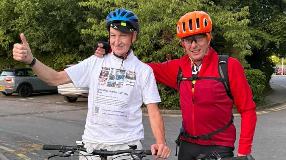 Two men standing astride bicycles.  One man is wearing a white t-shirt and a cycle helmet and is doing a thumbs up sign, a second man wearing a red fleece and cycle helmet has his arm around the first man.  They are standing in a carpark.