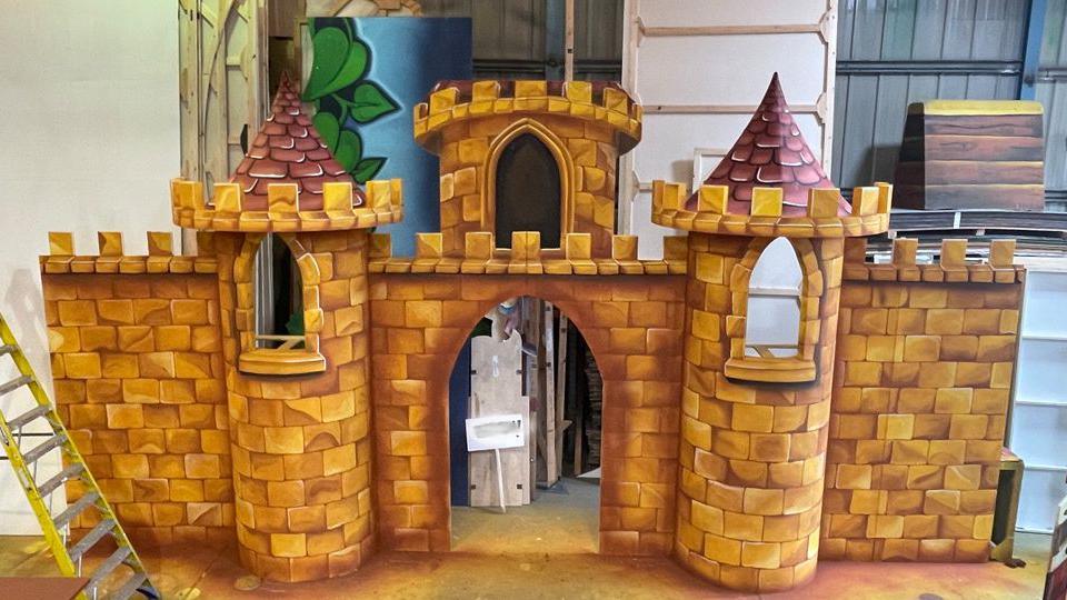 A pantomime set being built, it shows a castle, with brown bricks, purple turrets, and props around it. You can see other parts of sets behind it and a ladder to the left. 