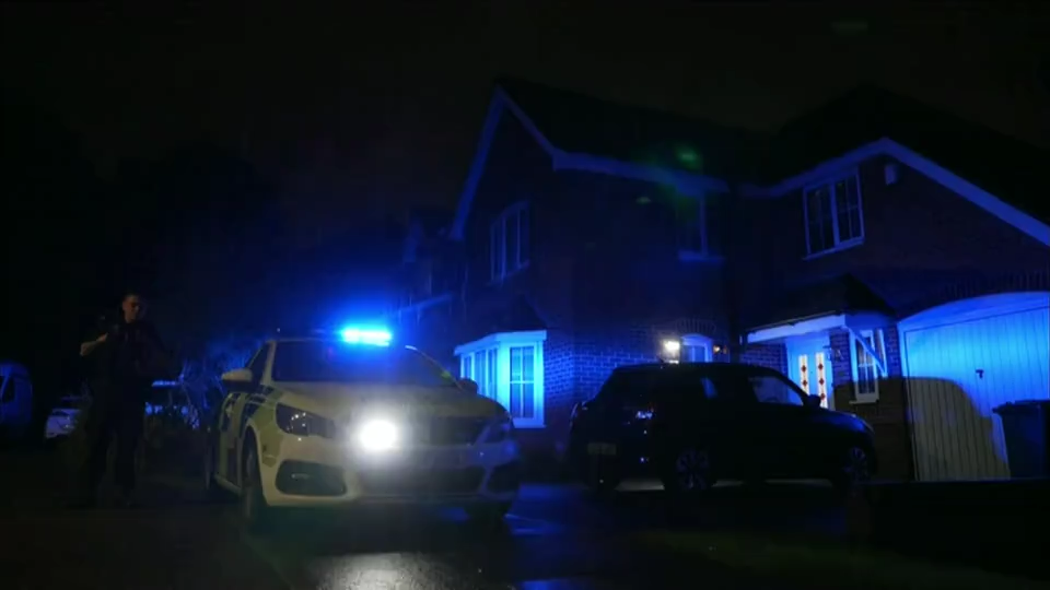 A police car is parked in front of a house with blue lights flashing. It is nighttime, so visibility is low, but an officer can be faintly seen standing next to the car