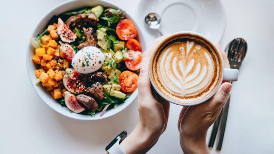 Hands holding a cup of coffee, colourful salad sitting on a white table next to the hands.