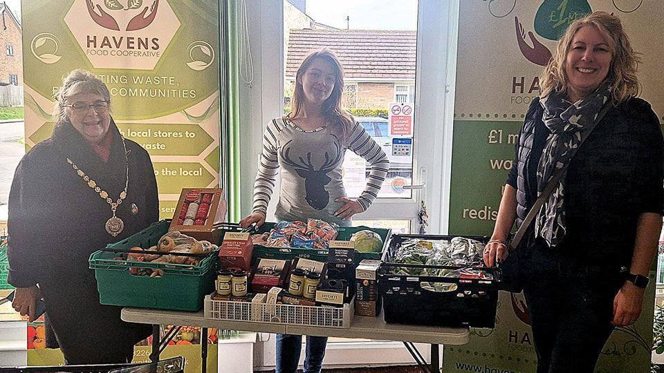 Three women standing behind baskets full of food.