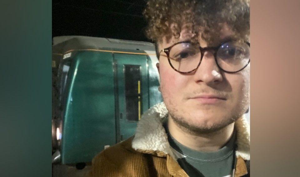 Marcus Patterson is standing in front of a green train. He is wearing glasses and the photo was taken in the dark. He has curly brown hair.