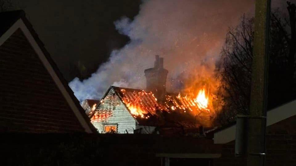 The pub with flames coming out of roof. The roof tiles are largely missing and the wooden beams remaining are on fire. Smoke is pouring into the night sky. 