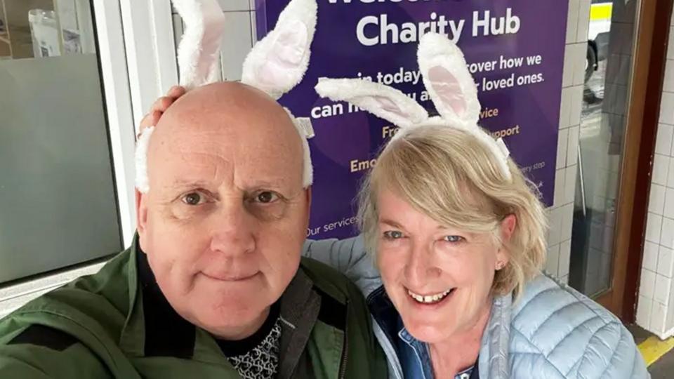 A man and a woman stand outside of a charity hub wearing white bunny ears on their heads.
