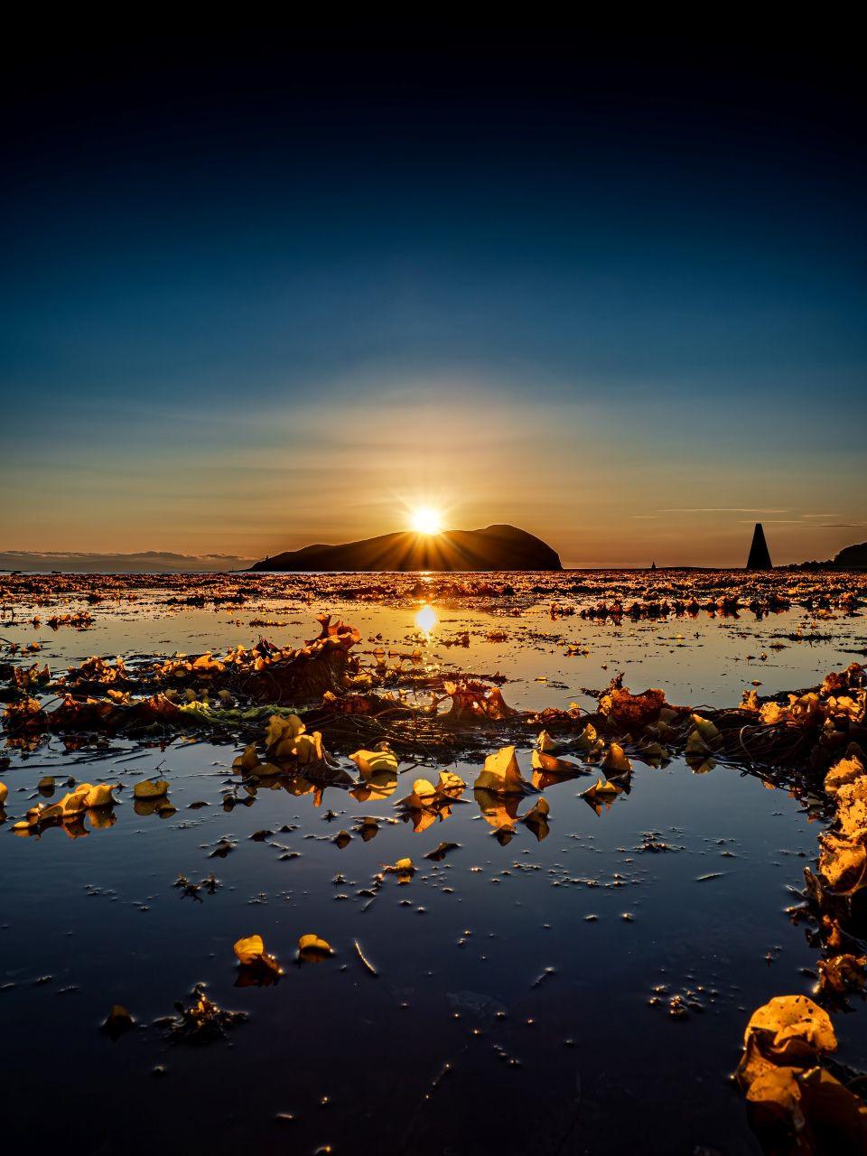 A sunset over an island with the sun illuminating rocks on a loch in the foreground