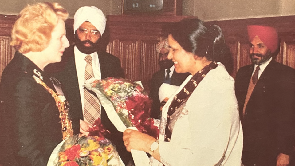 A woman presents flowers to Margaret Thatcher. Three Sikh men stand in the room.