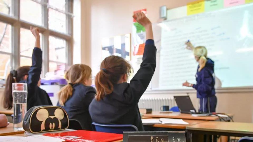 A teacher writes on a whiteboard at the front of the classroom. Three students wearing blue blazers watch on, two raise their hands as if t answer a question.