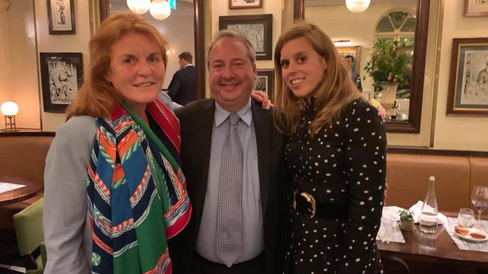 A photo of Jay Bloom from June 2019 in a restaurant with Sarah Ferguson on the left, wearing a pale blue top and a multi-coloured scarf, and Princess Beatrice on the right, wearing a navy dress with while polka dots and a belt with a large gold buckle.