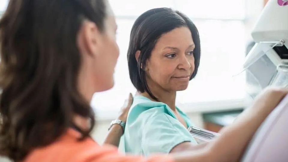 A woman wearing a great hospital gown is having a mammogram. She is pictured alongside a female medical professional.