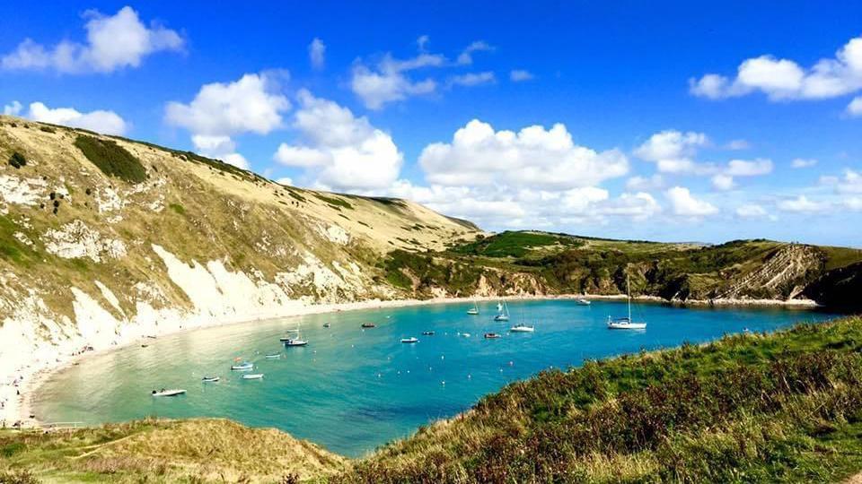 Seen from the cliff edge, several boats are in Lulworth Cove's green-blue water, surrounded by cliffs and with rolling fields in the background under blue skies