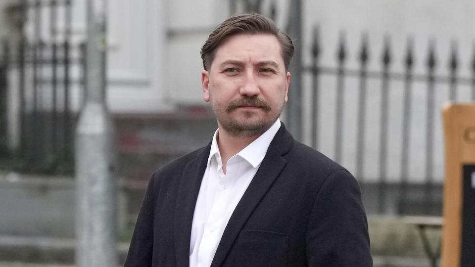 A man with a brown moustache wearing a white shirt and black jacket 