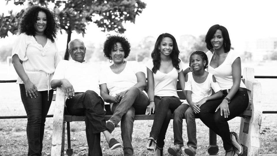 A black and white photograph showing smiling family members, from left to right: Melanie Clarke, Hugo Clarke (her father), Margaret Clarke (Melanie's mother), Danielle (Melanie's niece), Daelen (Melanie's niece) and Gillian (Melanie's sister). Melanie is standing beside a bench that everyone else is sitting on. They are all wearing light coloured tops and dark trousers or jeans. Picture taken in 2013