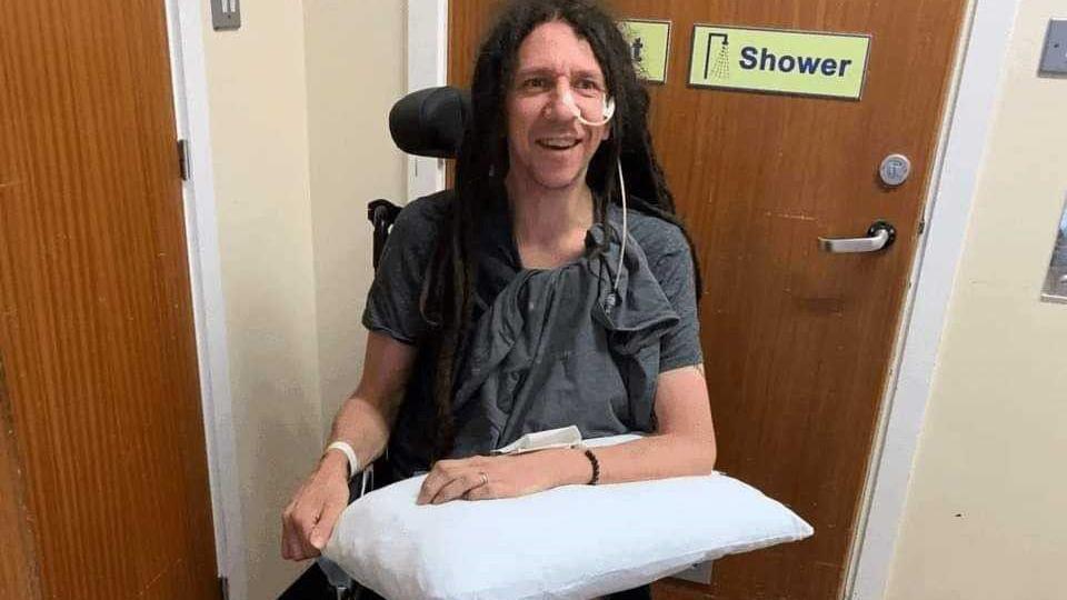 Tony sitting in a wheelchair in hospital, with a pillow on his lap and tubes attached to his face.