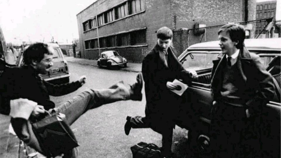 Grainy black-and-white photo of a young Bernard Sumner kicking his leg towards a young Ian Curtis. Rob Gretton is nearby, smiling and leaning against a car. They are standing in a street in front of a brick warehouse.