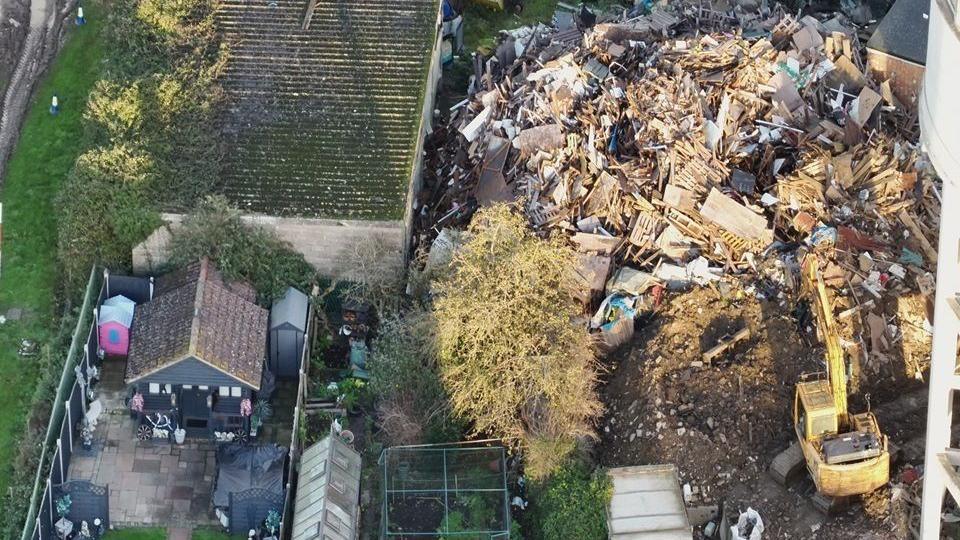 An aerial view of a skip yard with a large amount of wooden waste and soil. There is a yellow digger in the yard too. The yard is next door to a small garden, where there is a navy shed.