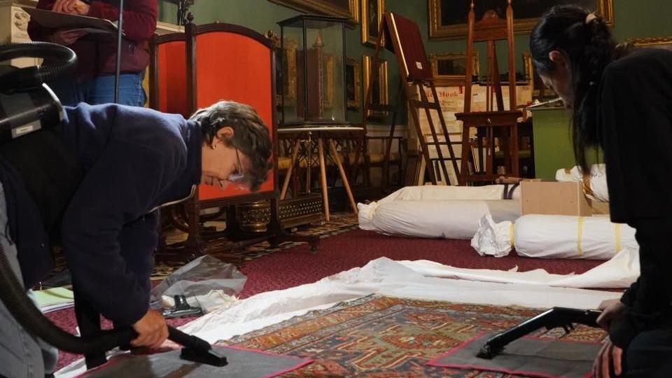 Two people in the room of a stately home surrounded by antique furniture bent over an antique rug with special mats and small hoovers.