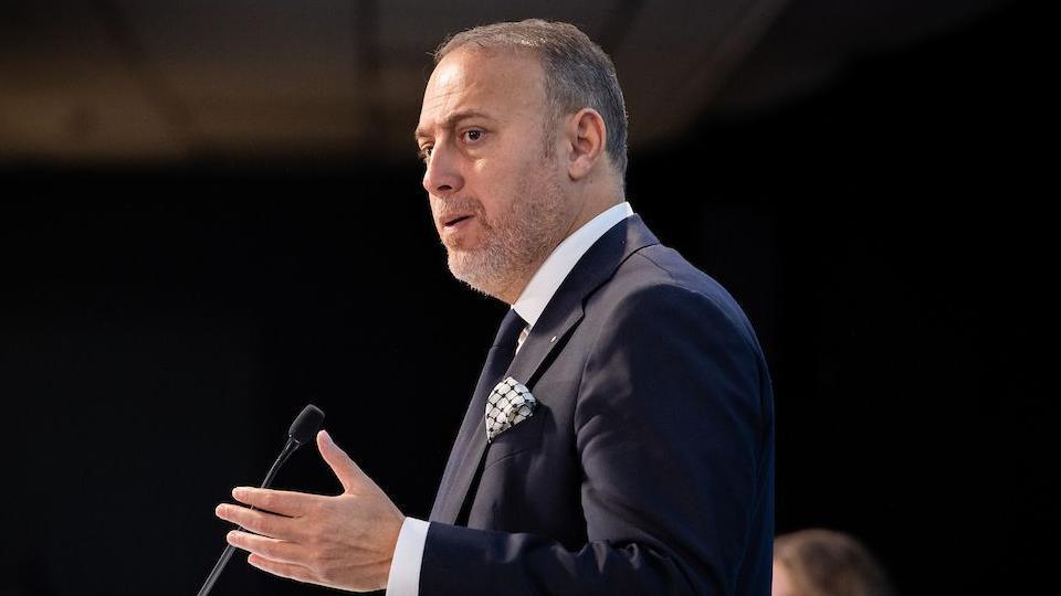 Husam Zomlot speaking at Plaid Cymru conference in front of a microphone, with a black tie and a black suit, and a white shirt.