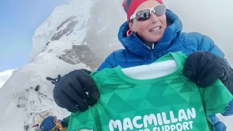 A woman in a blue coat and red hat and blue gloves, holding a Macmillan cancer support t-shirt in the mountains in Nepal.