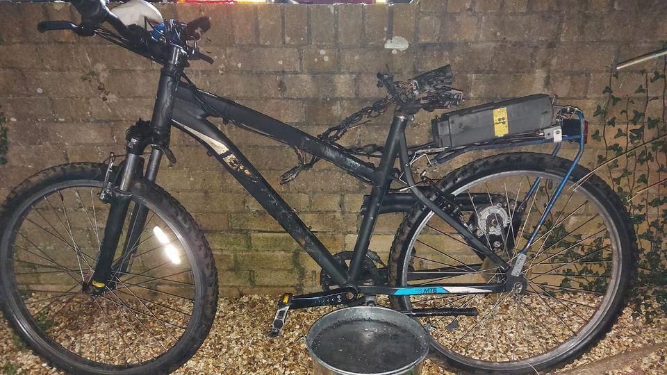 A blackened, burnt out bike that was modified with a cheap e-bike battery. There is a silver bucket of water in front of it.