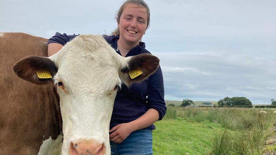 A young woman with black hair tied back is smiling at the camera with her arm around a brown and white cow with yellow tags in its ears. She has a navy zip-up fleece and blue jeans. Behind her is green field.