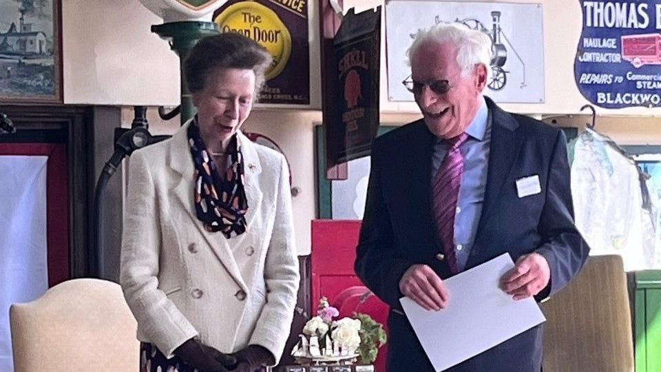 Princess Anne smiling with Malcolm Stern who is also holding and holding a certificate. They are on stage together at an event.