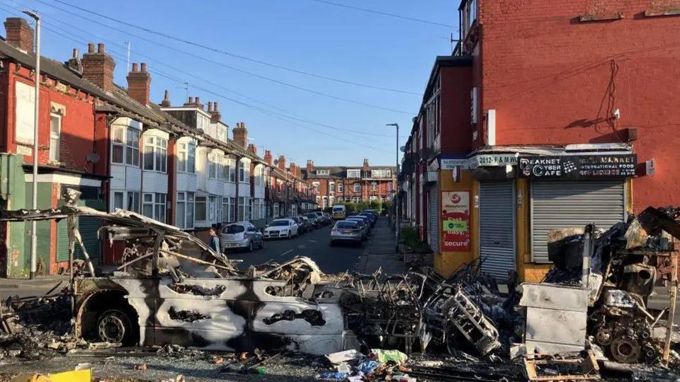 A street after a riot with a burnt-out bus and debris in the road 