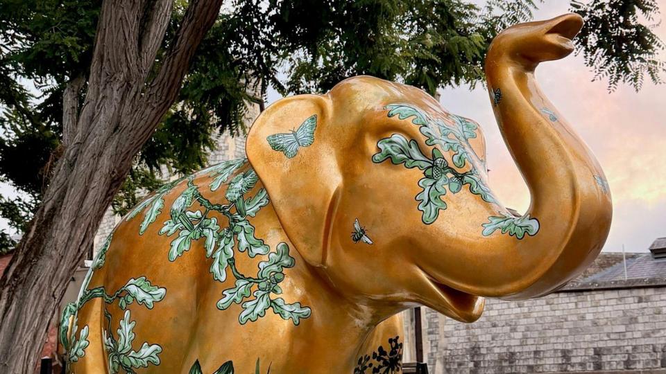 An elephant sculpture is pictured next to a tree. It is gold in colour with green leaves and foliage painted around its body. 