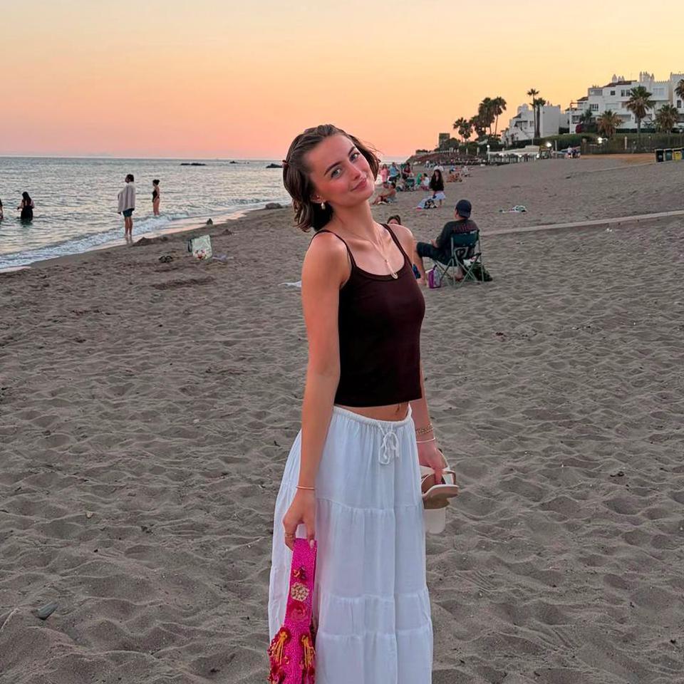 Person standing on a sandy beach at sunset, wearing a dark sleeveless top and a white skirt, holding sandals in one hand and a bright pink accessory in the other. The sea stretches along the shoreline, with several people near the water and white buildings and palm trees visible in the distance beneath a pastel orange and pink sky.